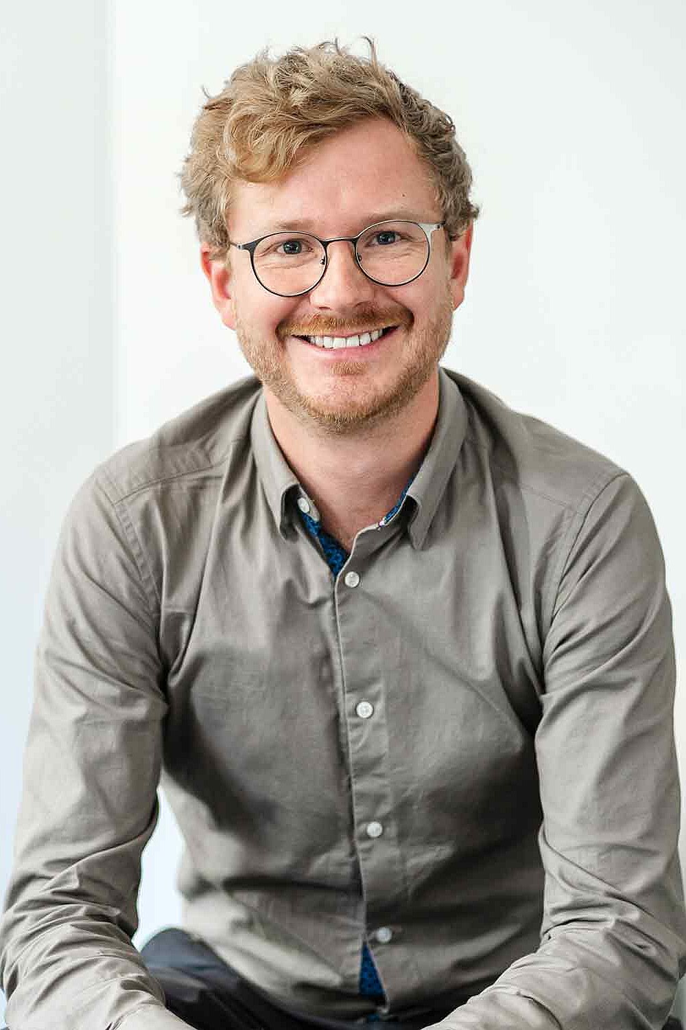 Johannes Walbrunn, Prokurist bei re-sult, hat blonde, kurze Locken, trägt eine Brille und sitzt locker auf einem Stuhl, leicht nach vorne gelehnt. Er lächelt herzlich in die Kamera.